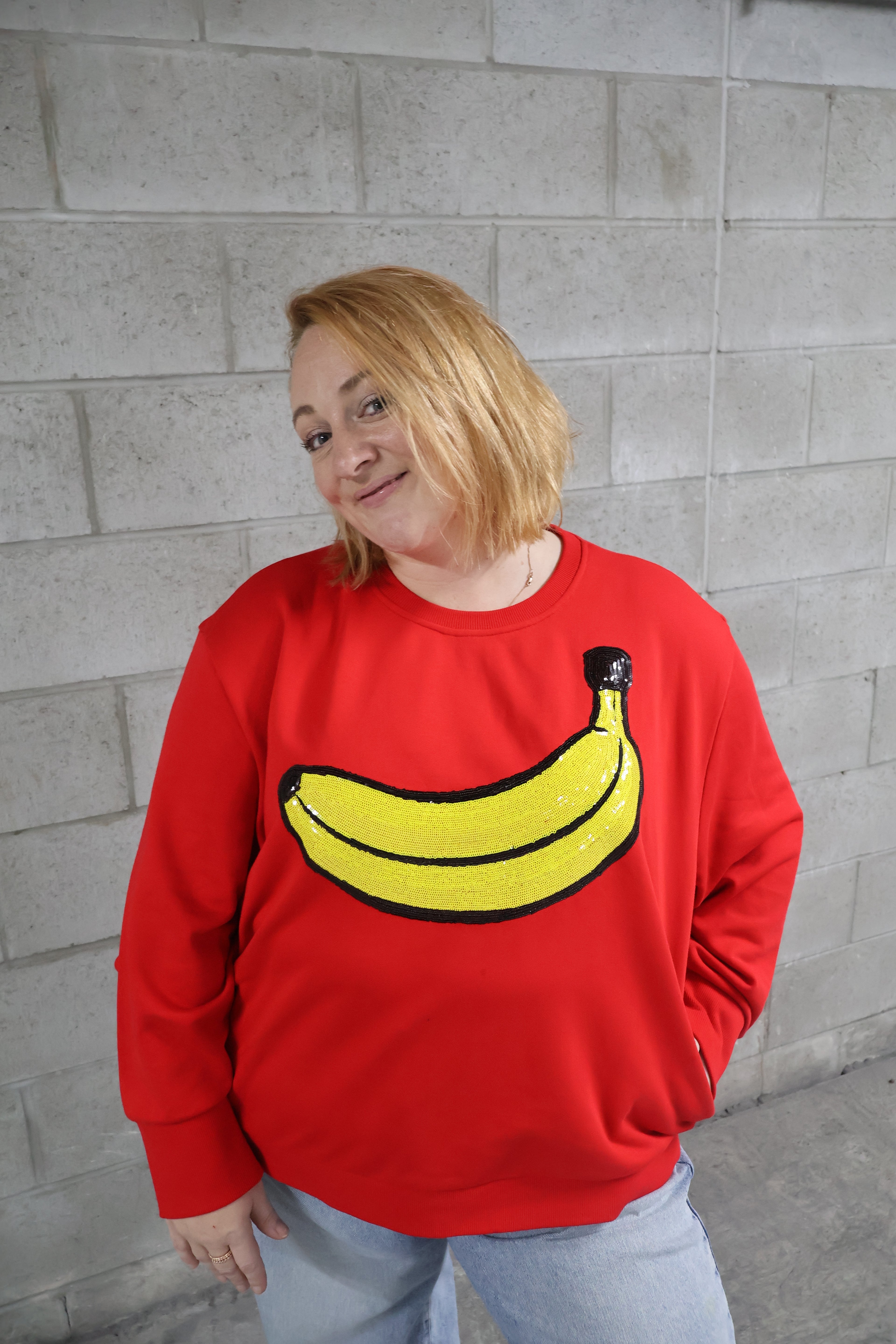 Person wearing a red sweatshirt with a banana design against a gray brick wall.