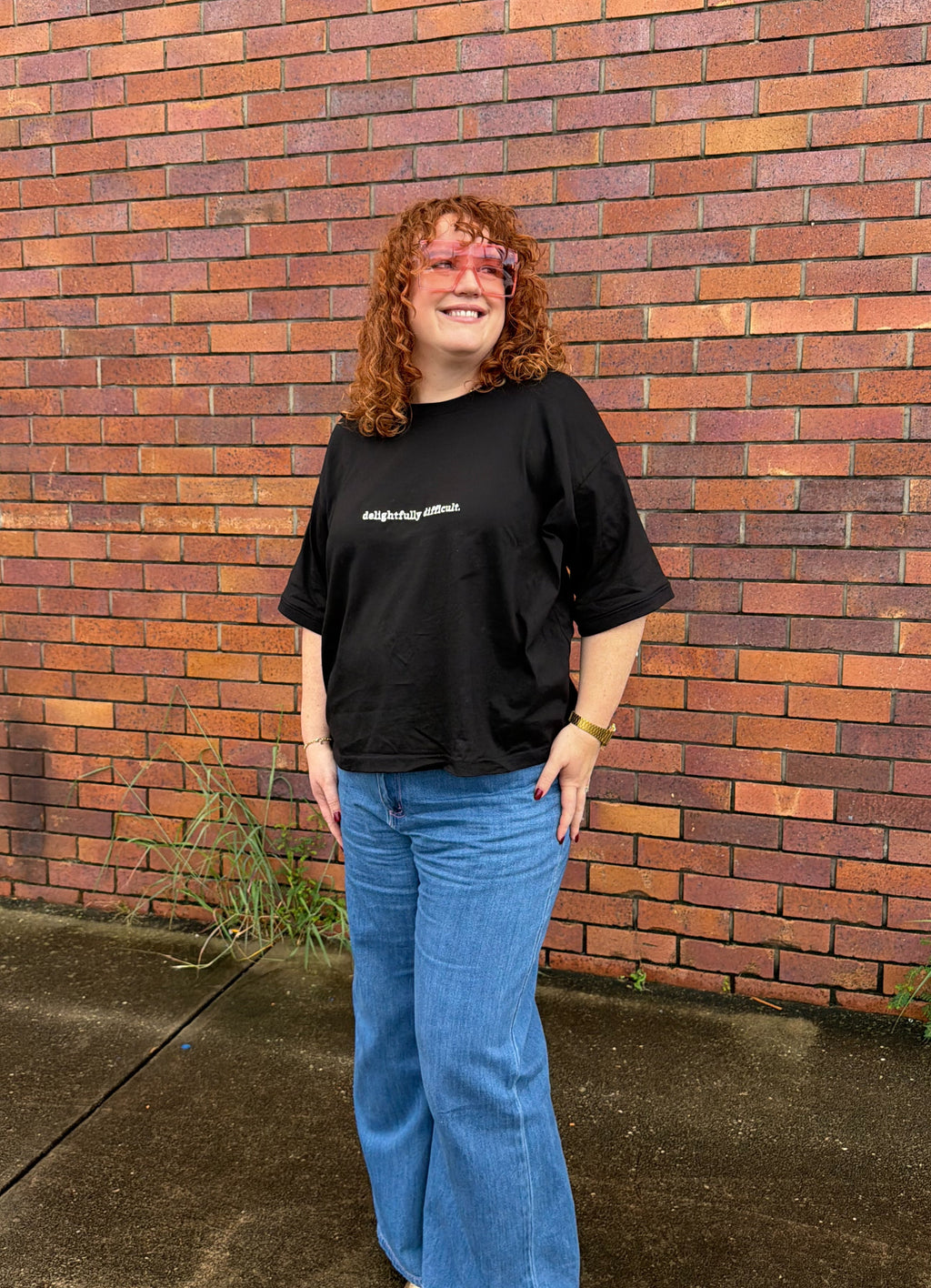 Person wearing a black t-shirt and blue jeans standing in front of a brick wall.