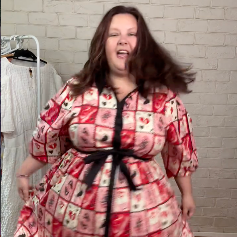 Person wearing a patterned dress with a black belt in a room with a brick wall.