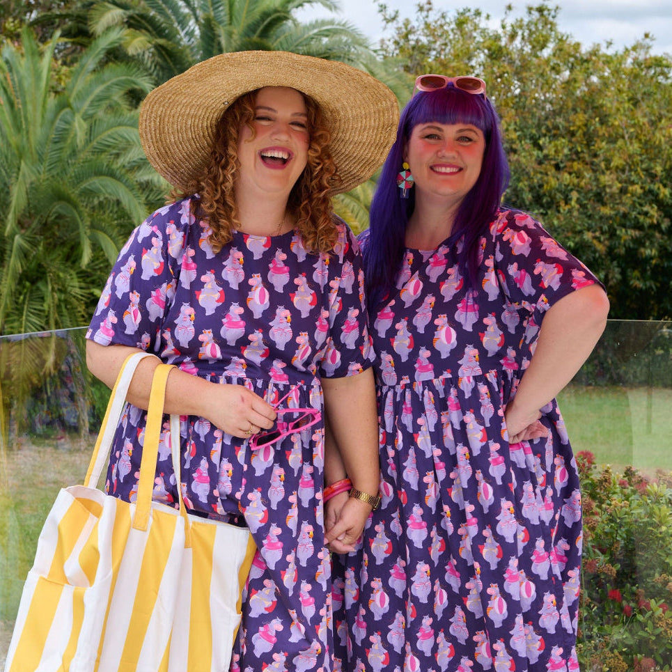 Two women wearing matching purple dresses with a hippo pattern, standing outdoors.