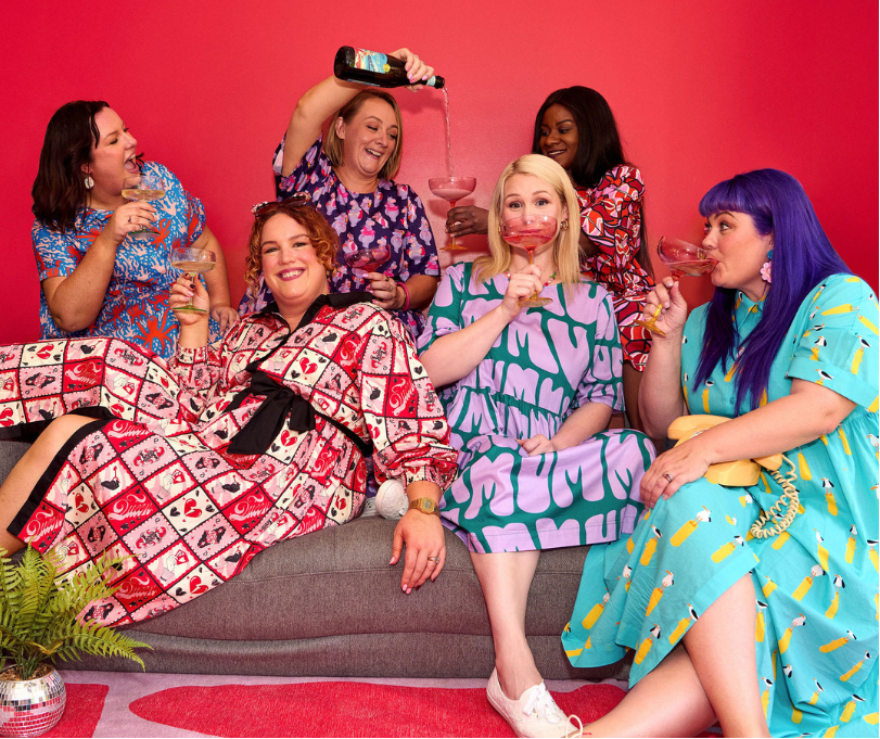 Five women sitting on a couch against a red wall, with a pink accent at the top.
