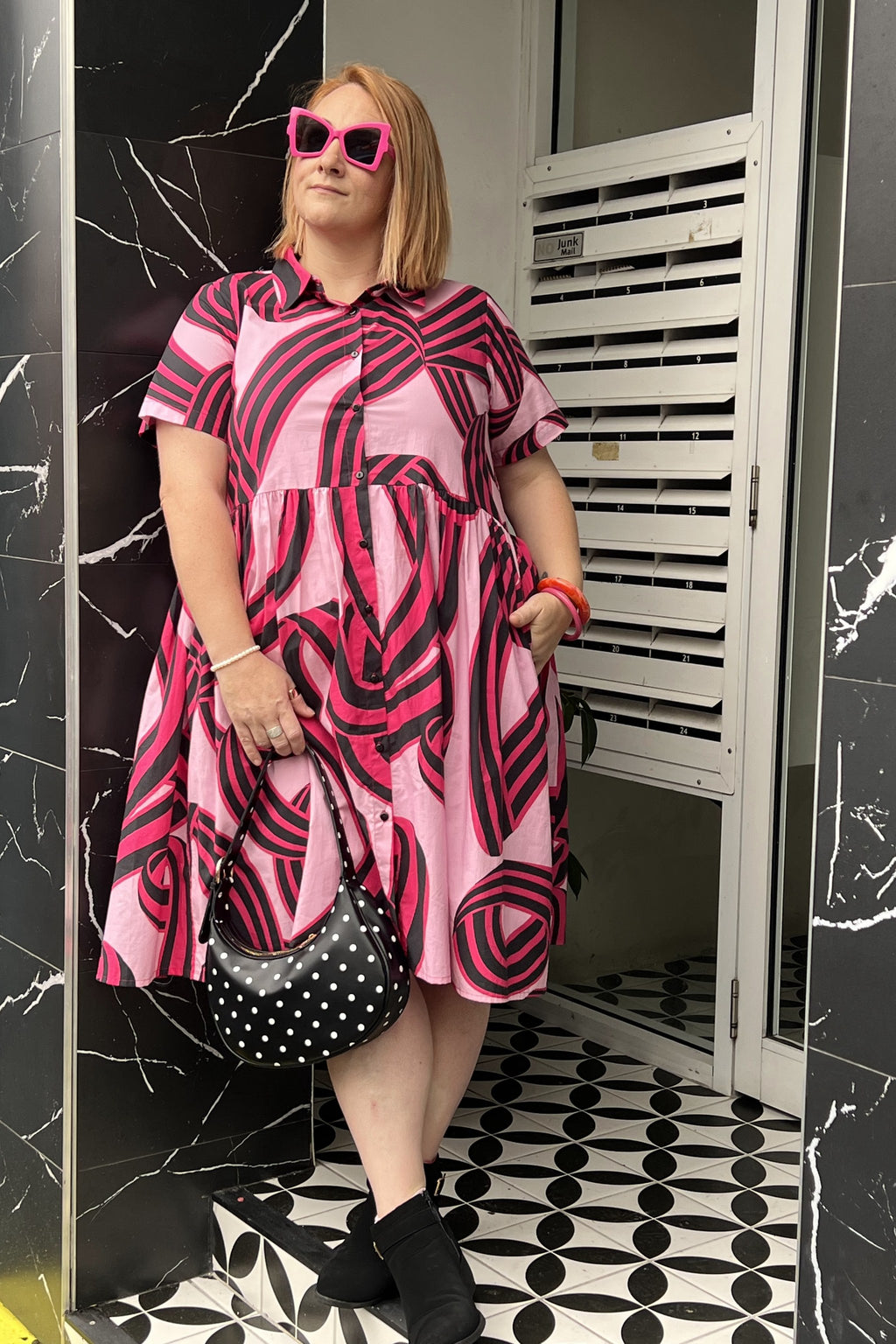 Toomi Liquorice Swirl dress, full length, worn with pink sunglasses and a polka dot bag against black marble