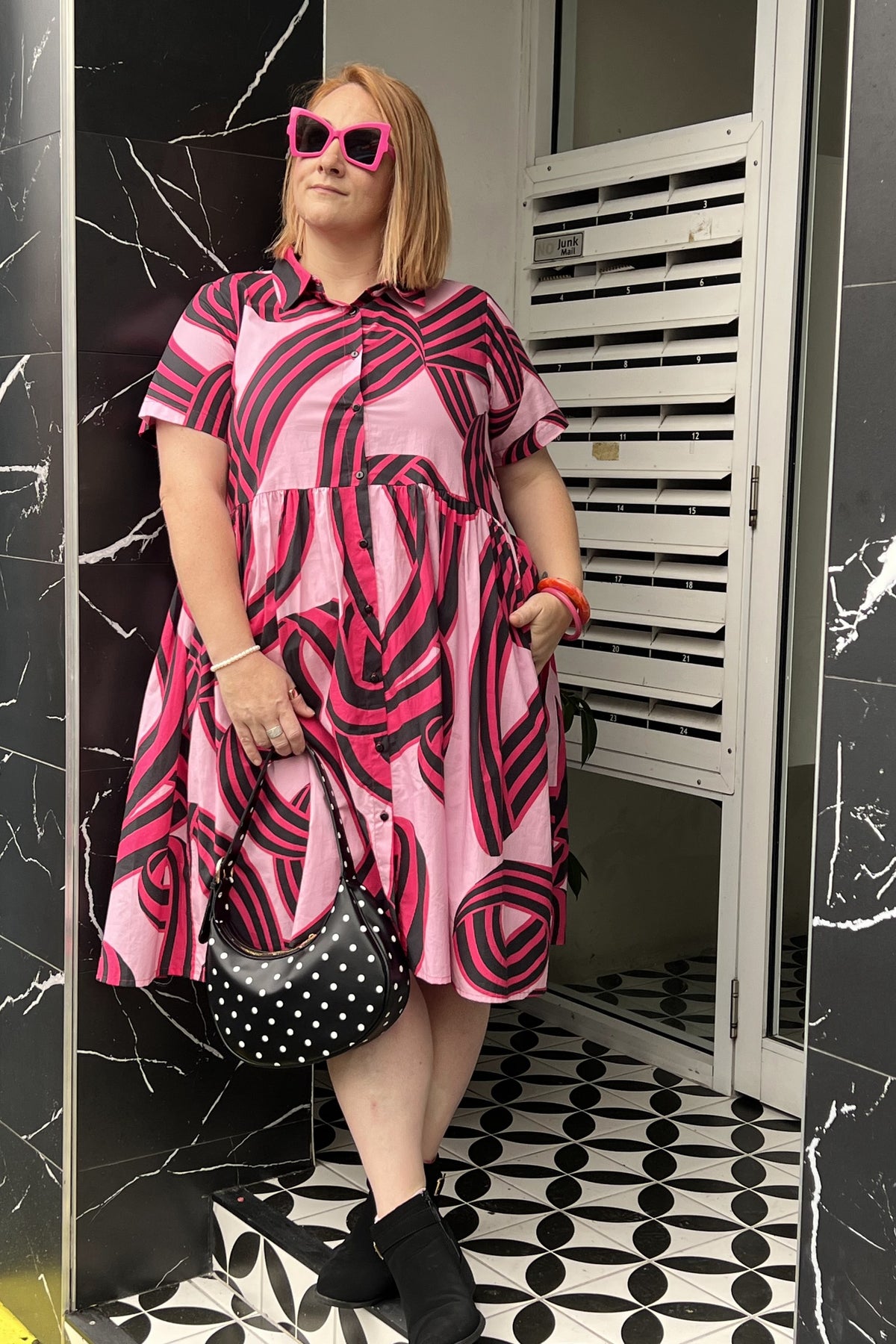 Toomi Liquorice Swirl dress, full length, worn with pink sunglasses and a polka dot bag against black marble